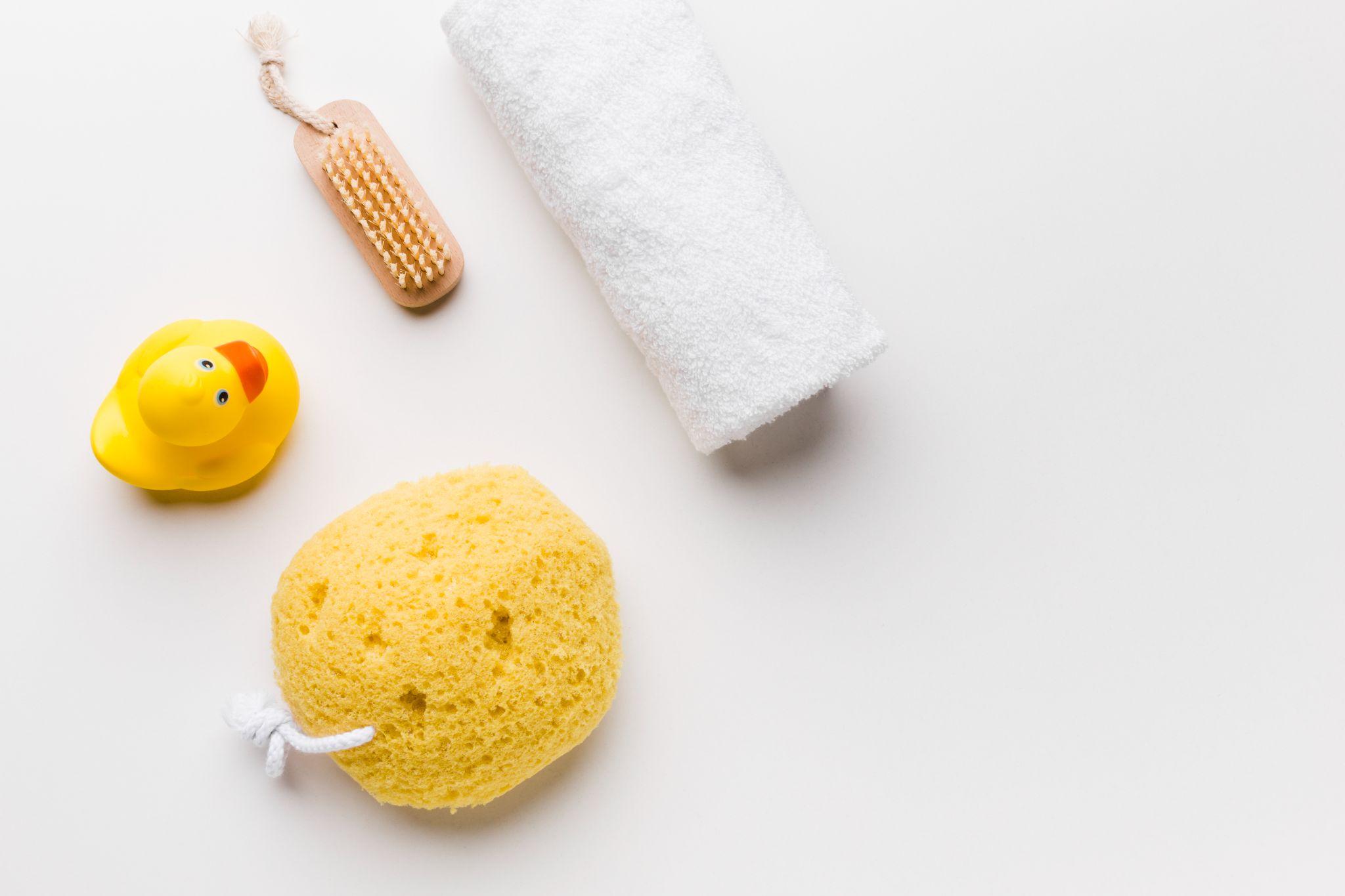 Na imagem, há uma esponja-de-banho, toalha, escova e um patinho de borracha espalhados em uma superfície branca.